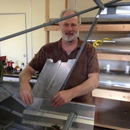 A man with a beard is standing in front of a machine holding a piece of metal.