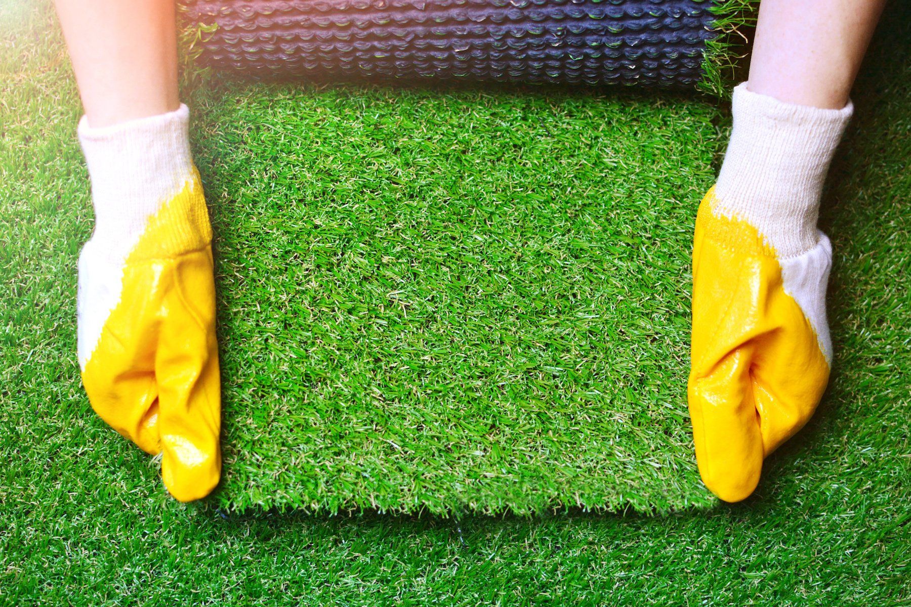 Hand wearing gloves carefully holding a piece of artificial turf against a lush green synthetic grass backdrop.