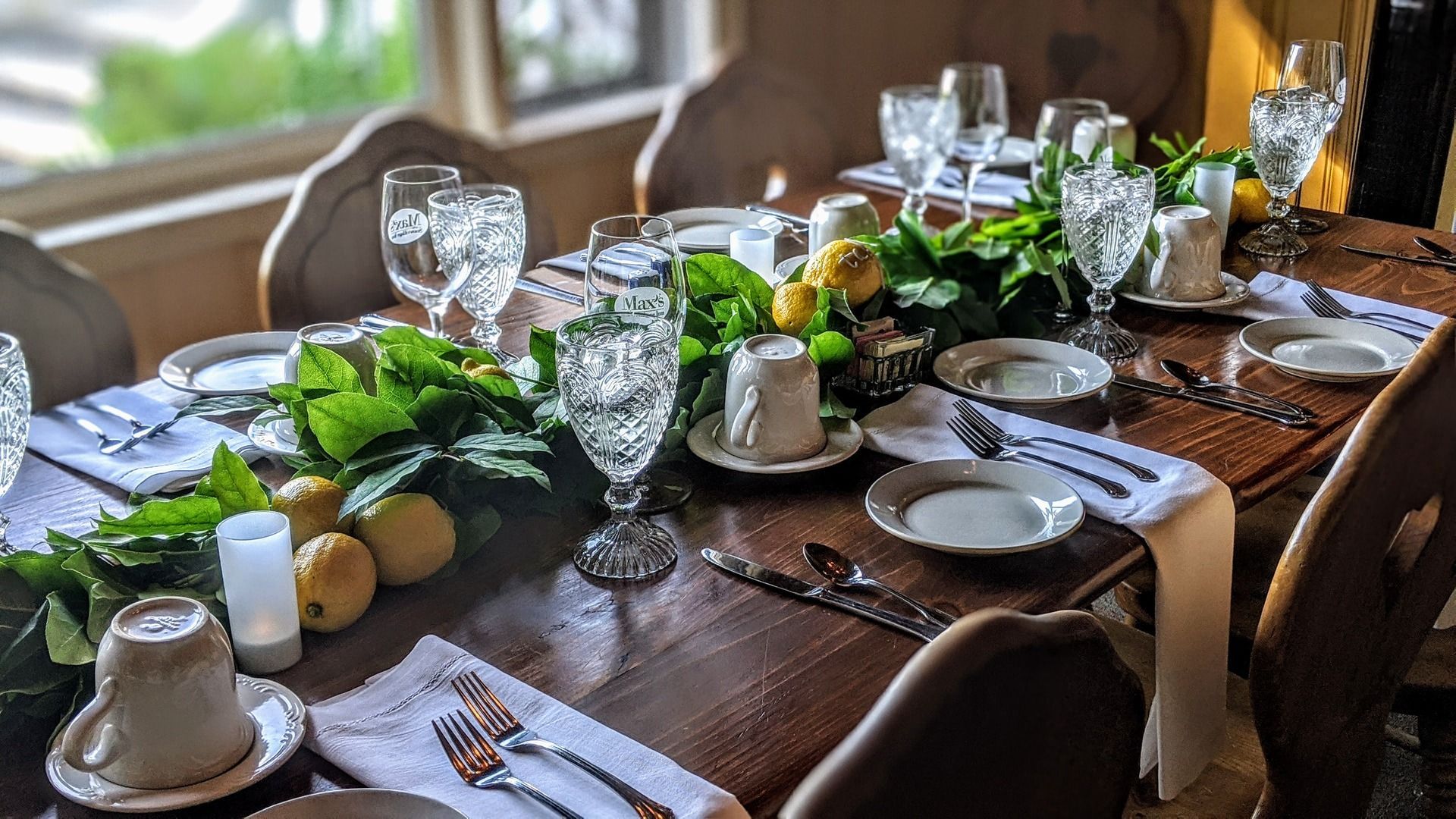 A long wooden table is set for a dinner party with plates , cups , glasses and utensils.