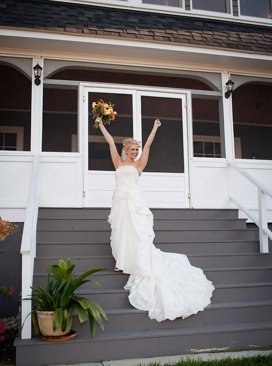 Bride in white dress, arms raised, celebrating on gray porch steps, holding bouquet.