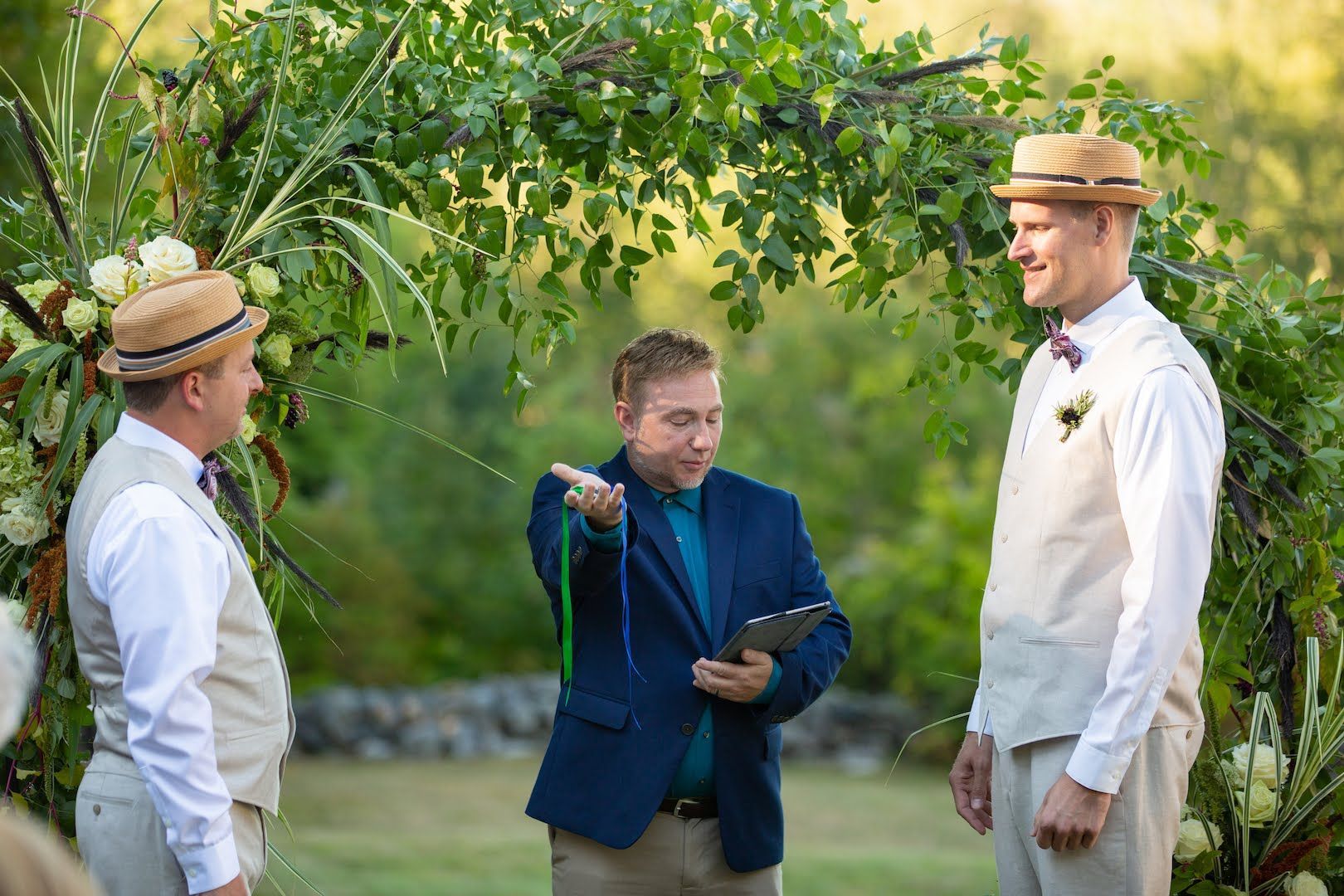 Three men at an outdoor wedding ceremony; one officiating, two wearing straw hats, standing under a greenery arch.