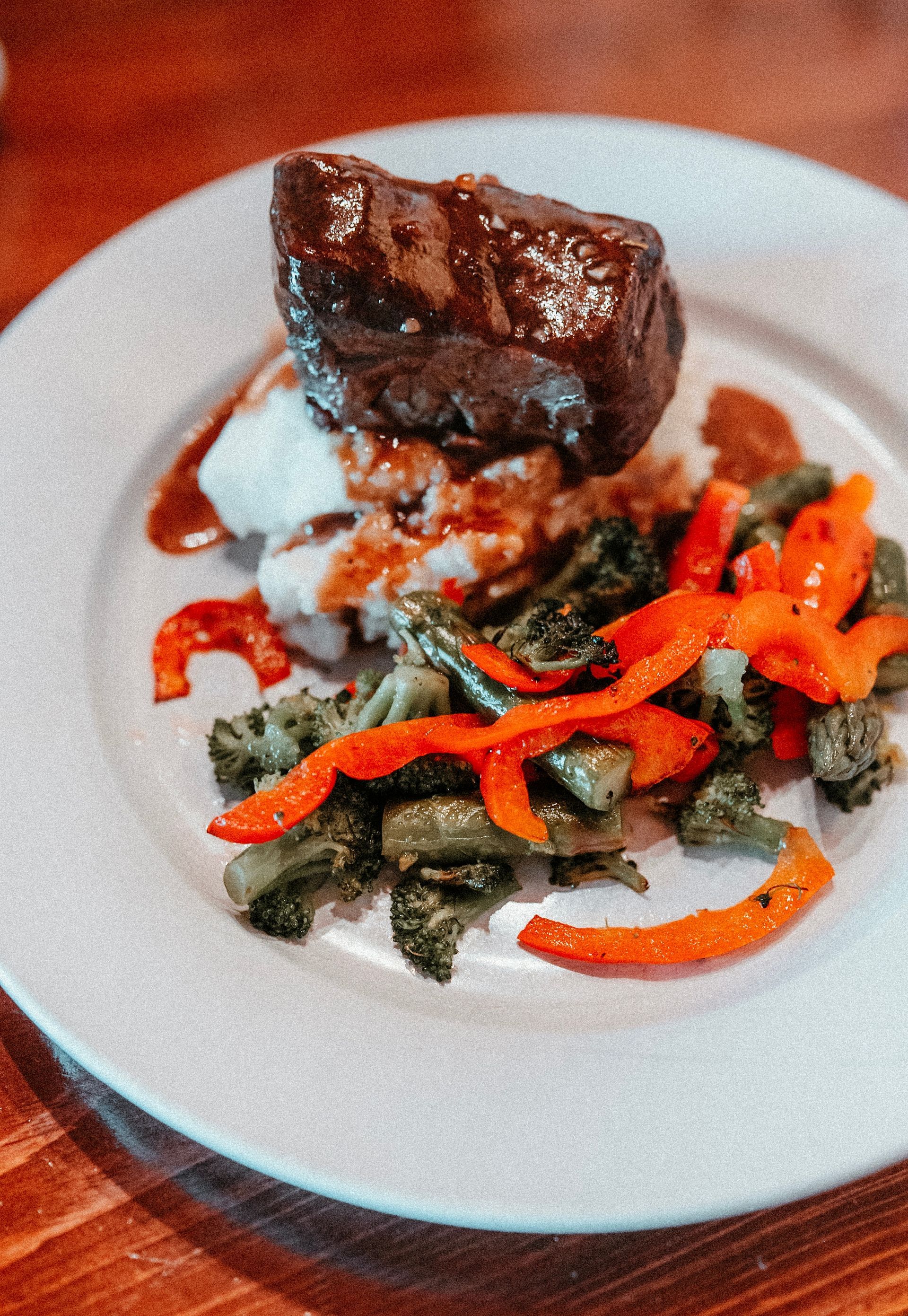 Steak, mashed potatoes, and roasted vegetables on a white plate. Dark meat with red and green vegetables.