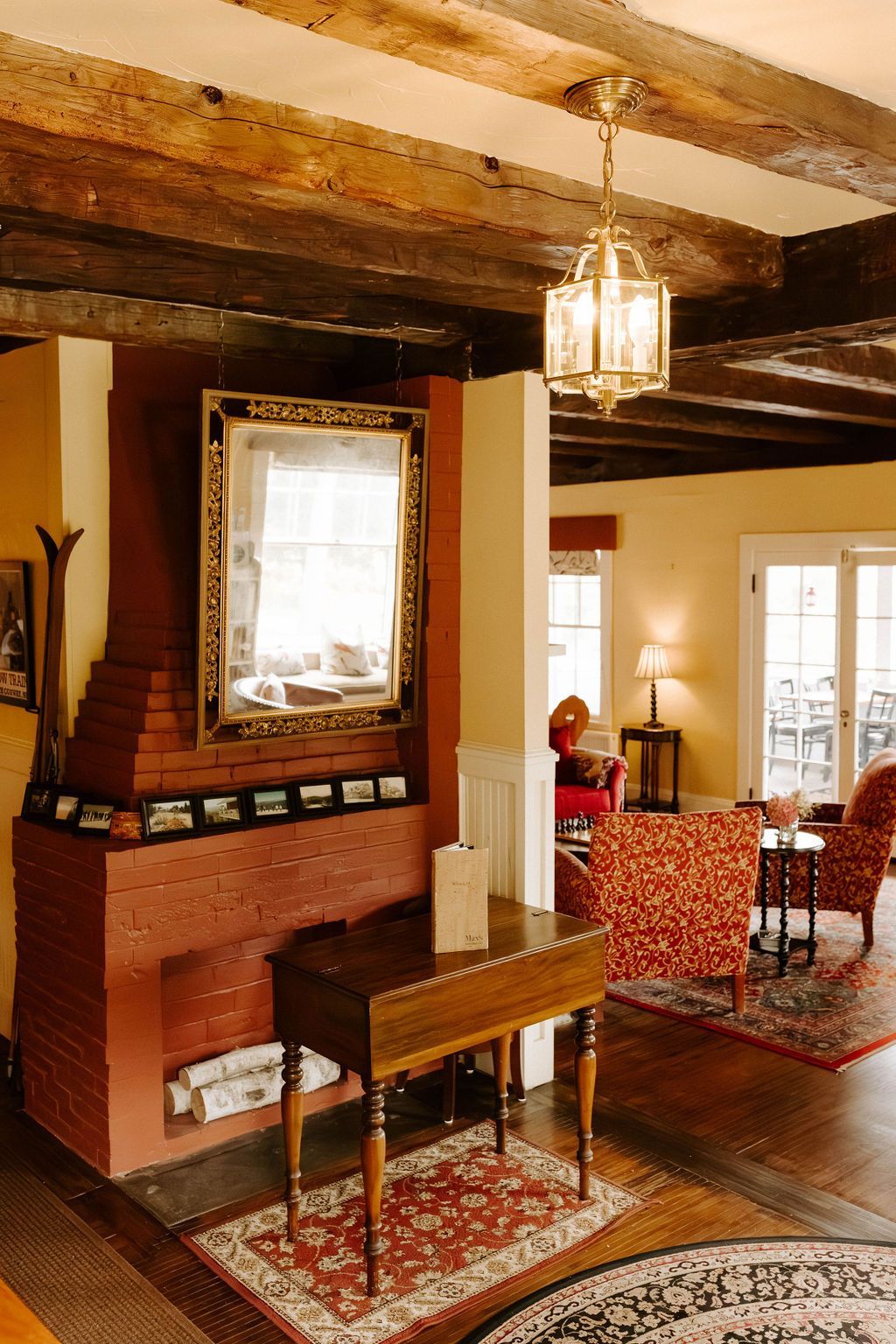 Cozy interior with exposed beams, brick fireplace, mirror, and two patterned armchairs.