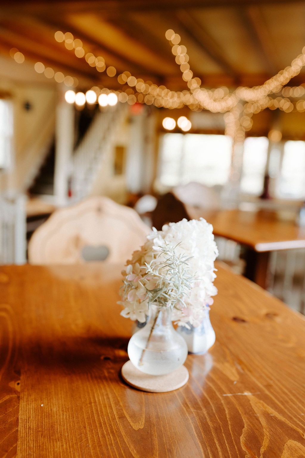 Small white flowers in a glass vase on a wooden table, in a restaurant setting with string lights.