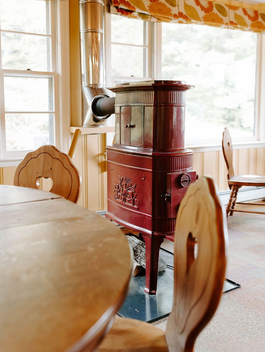 Red wood-burning stove in a cozy room with wooden chairs and a table. Windows provide natural light.