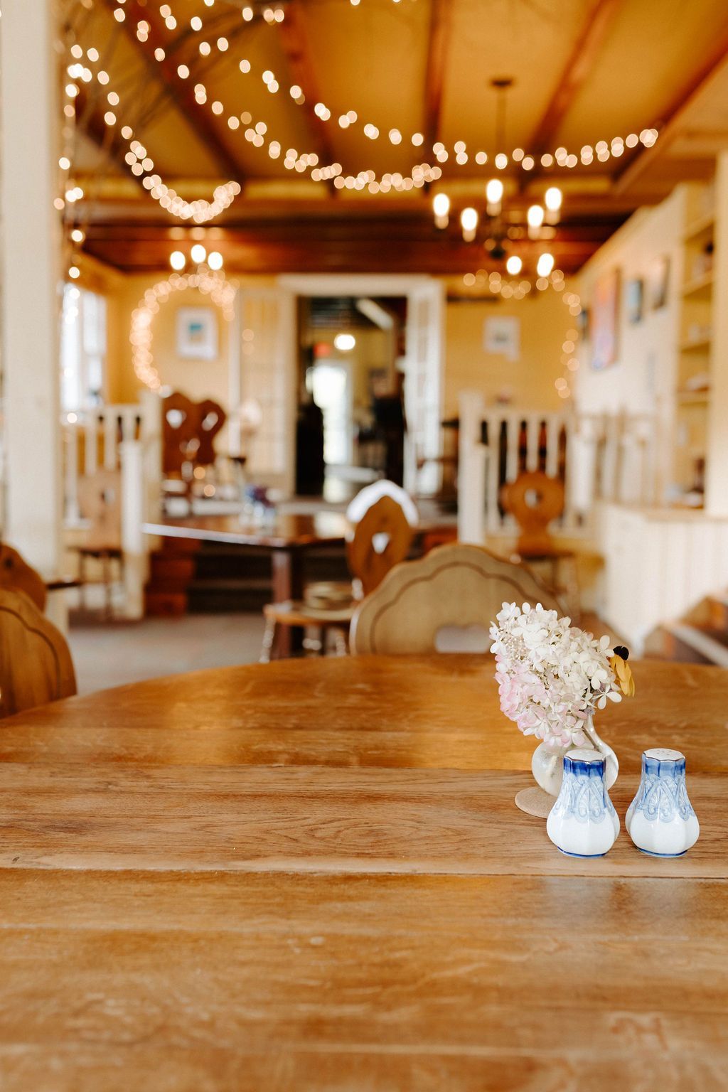 Wooden table in a rustic restaurant setting; string lights, decorative chairs, salt & pepper shakers, and flowers.