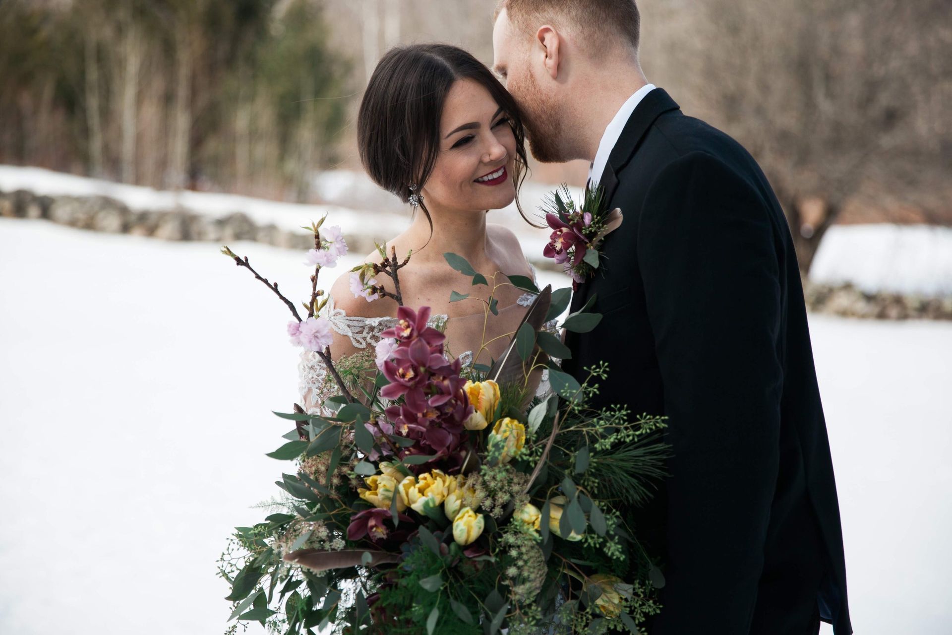Bride and groom embracing outdoors in a snowy landscape. The groom whispers, the bride smiles; she holds a colorful bouquet.