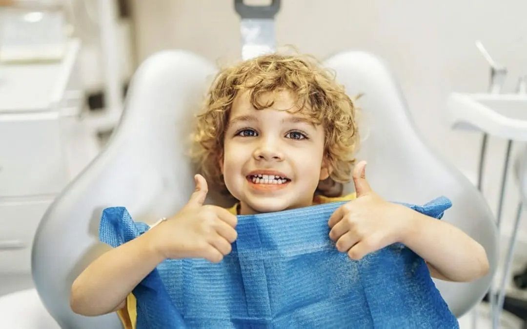 Boy with curly hair in a dentist's chair gives two thumbs up, smiling.