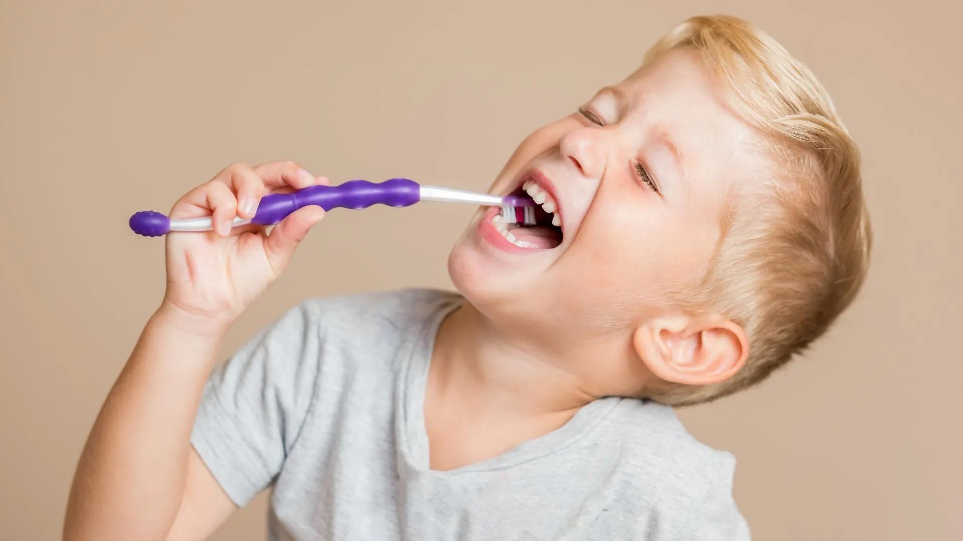 Young child brushes teeth with a purple toothbrush, eyes closed, smiling, against a neutral background.