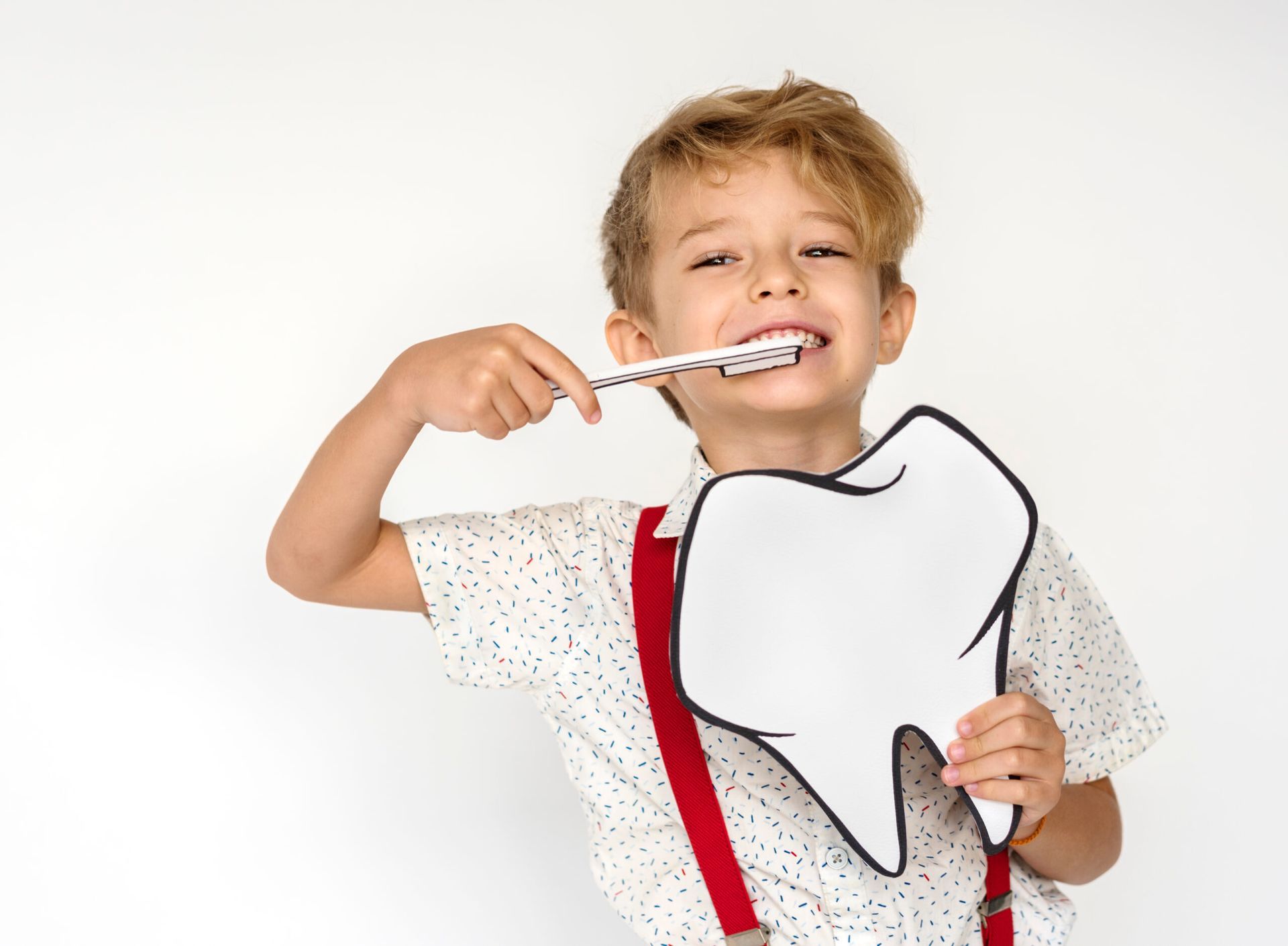 Young boy brushing a large tooth cutout, smiling. He wears a polka-dot shirt and red suspenders.