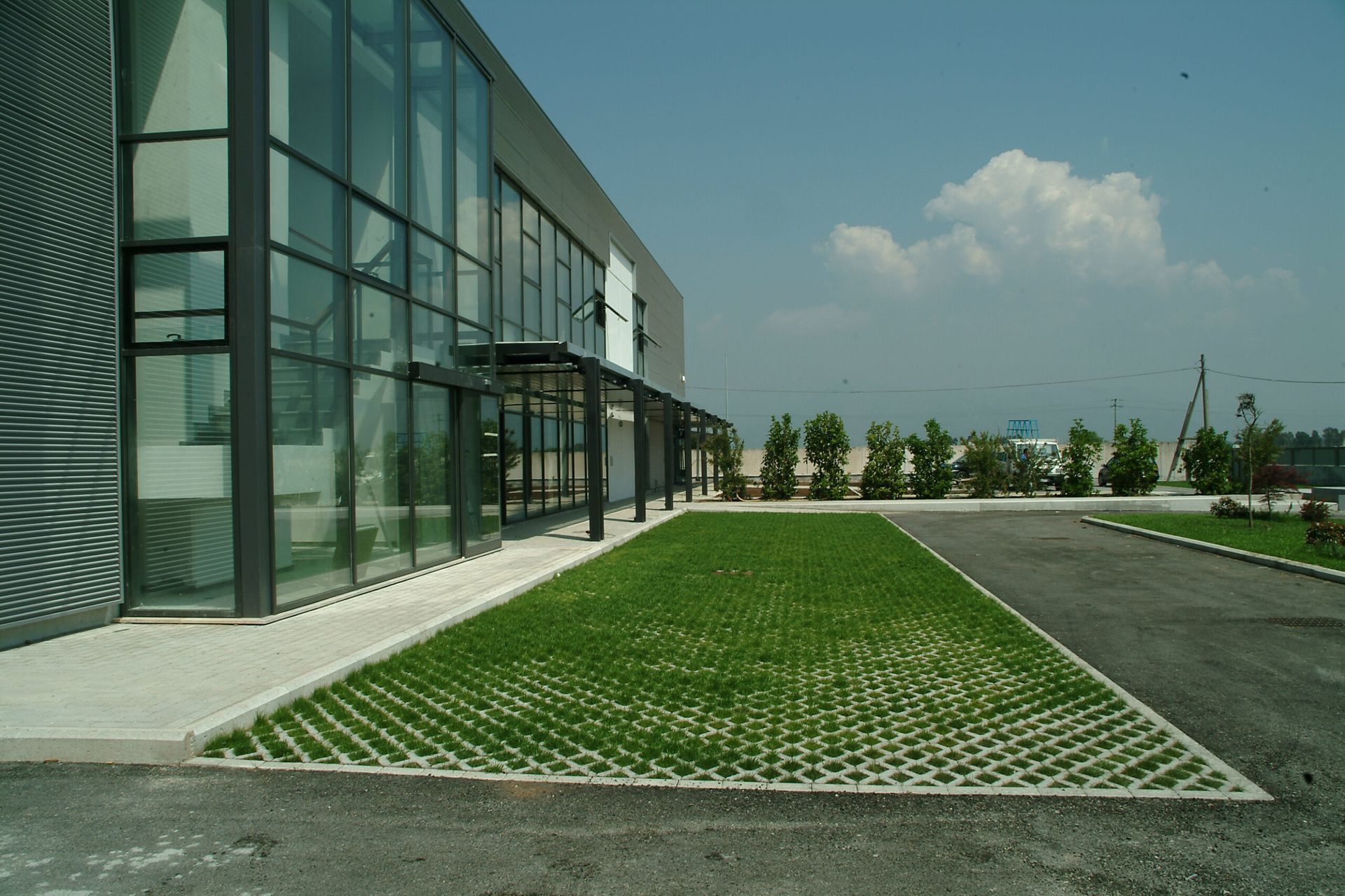 Edificio moderno con ampie finestre e un prato verde accanto a un parcheggio sotto un cielo azzurro.