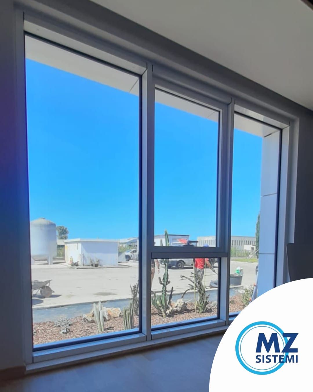 Una finestra luminosa mostra un cielo azzurro. Moderna finestra a triplo vetro con telaio grigio chiaro. In basso a destra è visibile il logo aziendale Mz Sistemi Latina