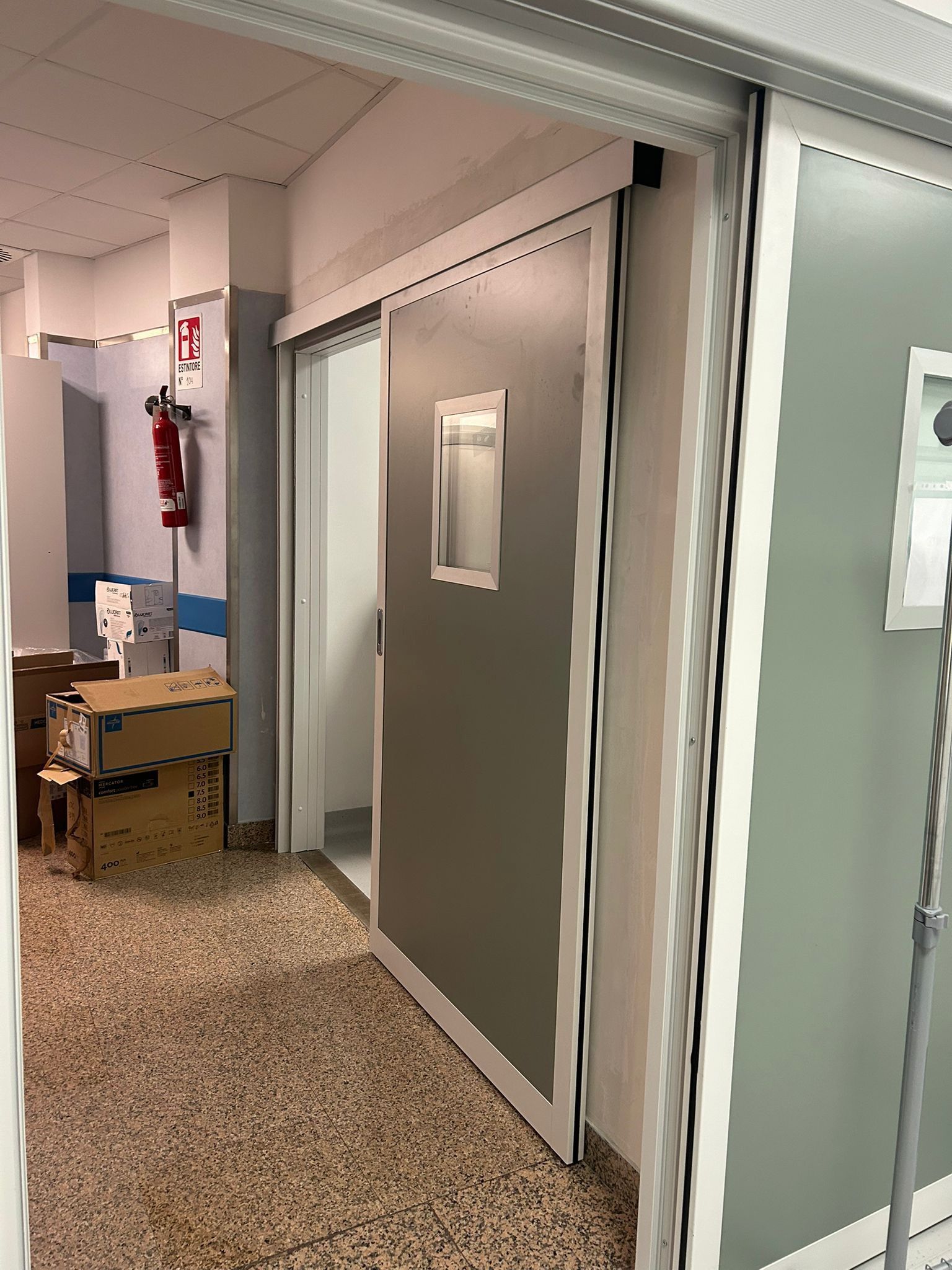 Corridoio dell'ospedale con porta scorrevole e una piccola finestra. Un estintore appeso al muro Mz Sistemi a Latina