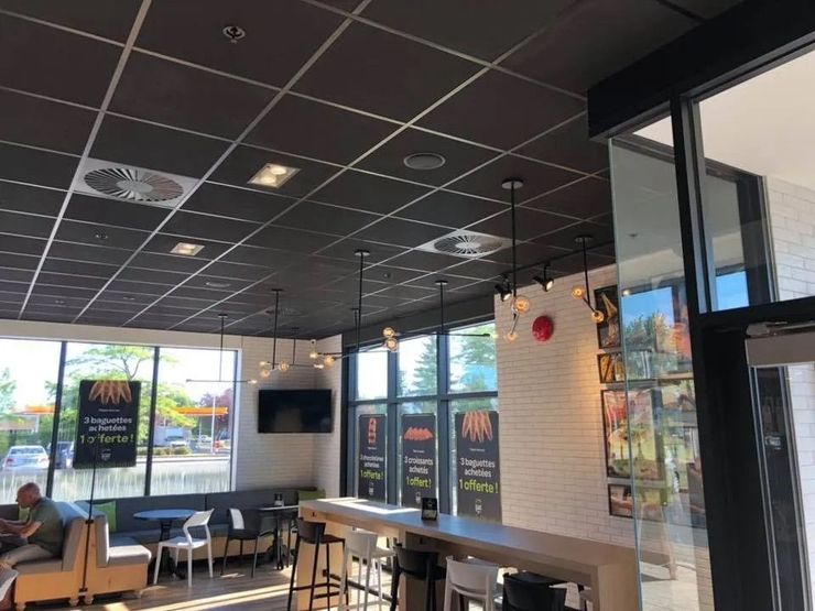 Un espace café intérieur moderne et lumineux avec un plafond à grille noire, des tables hautes, des tabourets, une vue sur l'extérieur et des affiches au mur.