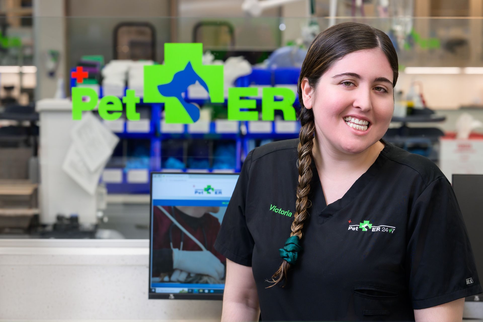Veterinary exam room: stainless steel table, surgical light, cabinets, window, gray floors, black chair, and stool.