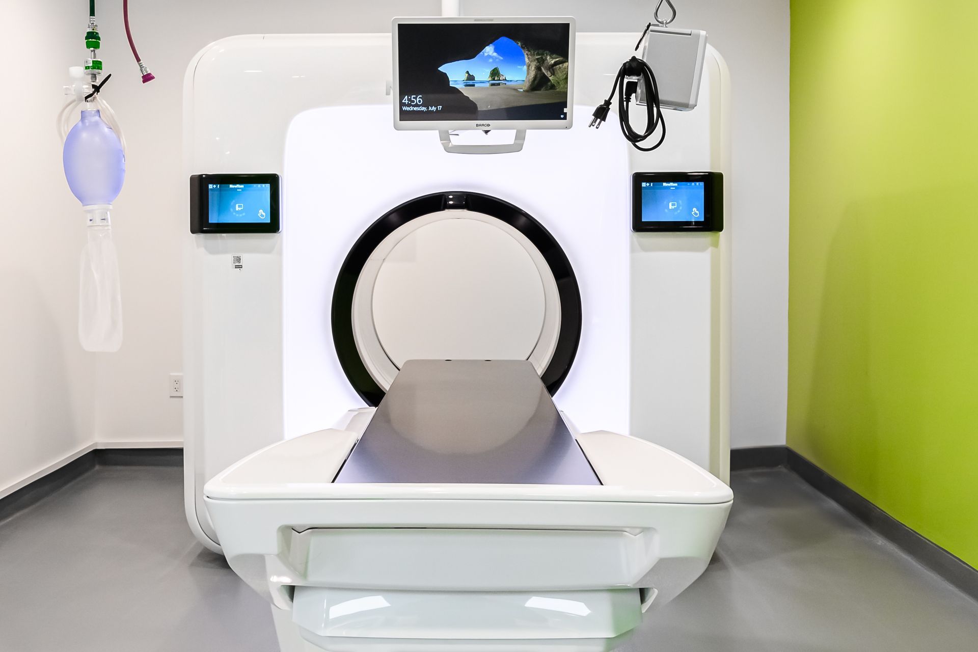 CT scan machine with a patient table, monitors, and medical equipment in a brightly lit room.