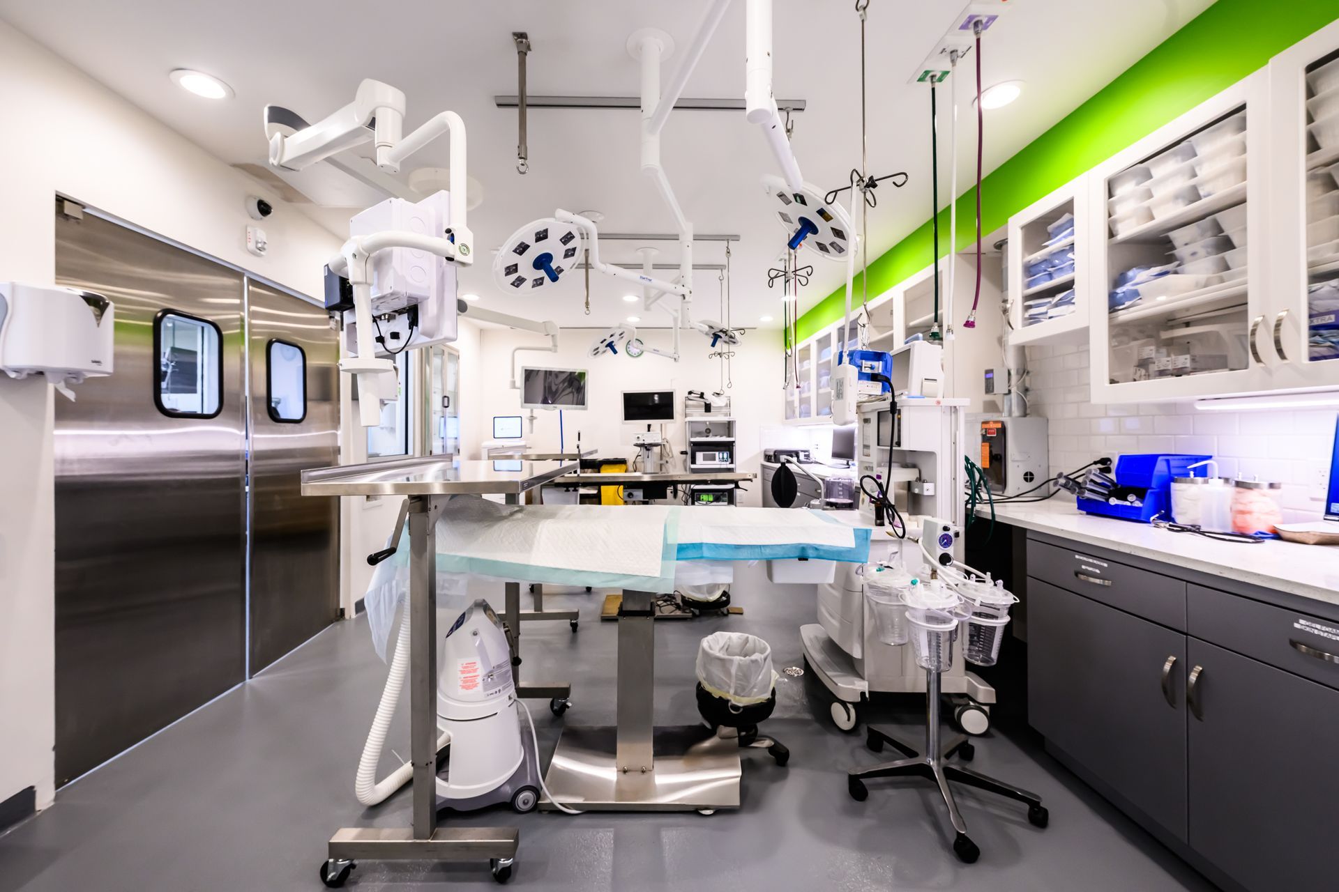 Surgical suite: stainless steel door, operating table, medical equipment, bright lights, white cabinets, green wall.
