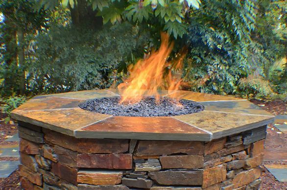 A lit, octagonal fire pit made of stone with a slate top, flames rising against a backdrop of green foliage.