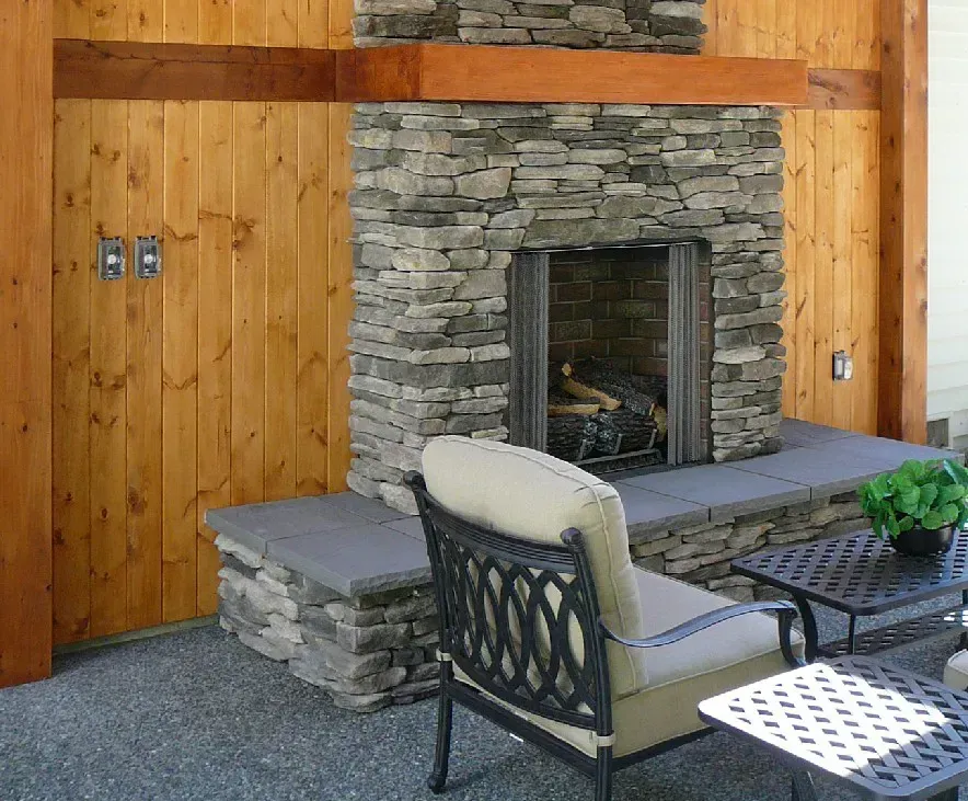 Outdoor stone fireplace with a wooden surround, next to a patio seating area. A gray armchair is in front of the fireplace.