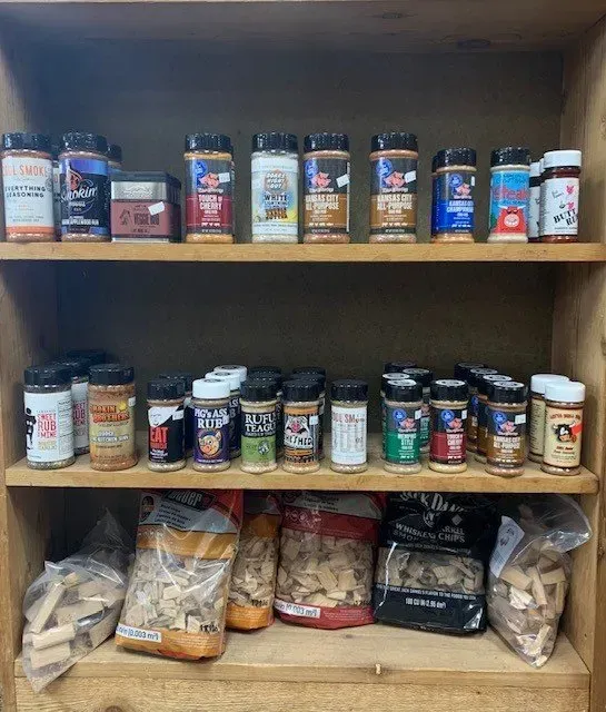 A wooden shelf displays various spice bottles and bags of wood chips for smoking, likely in a store.