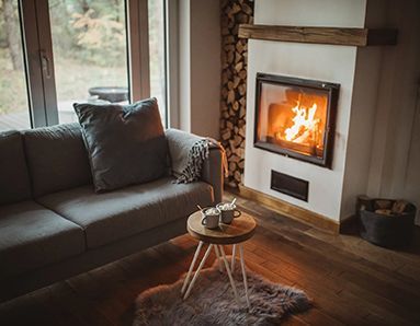 Cozy living room with a lit fireplace, grey couch, and small table with drinks. Wood flooring, logs stacked beside the fireplace.