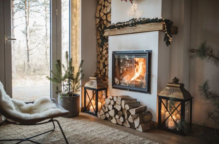 Cozy living room with a lit fireplace, lanterns, firewood, and a chair. A fir tree and a window with a forest view are also present.