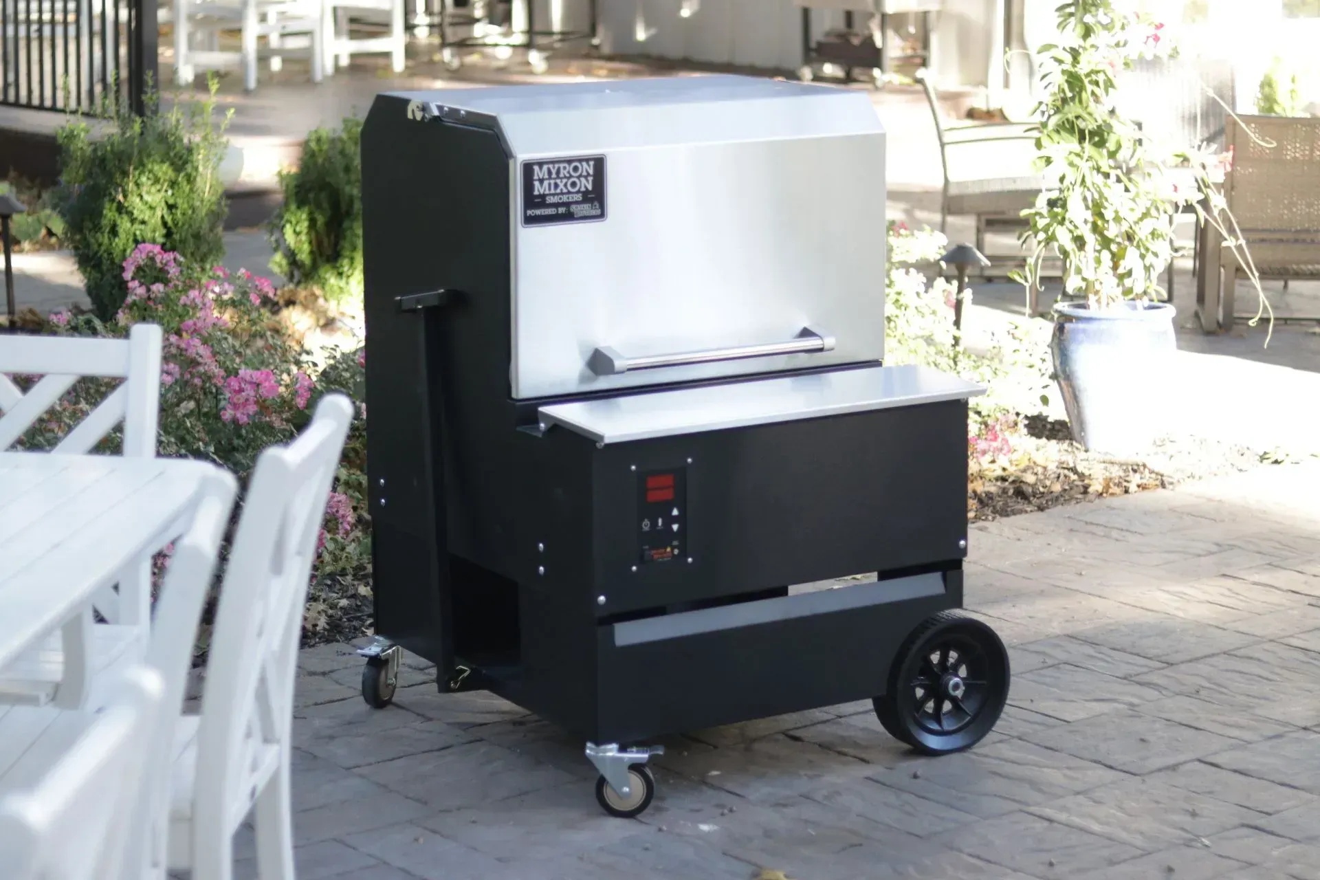 A black and silver smoker grill with wheels on a patio, near white furniture and flowers.