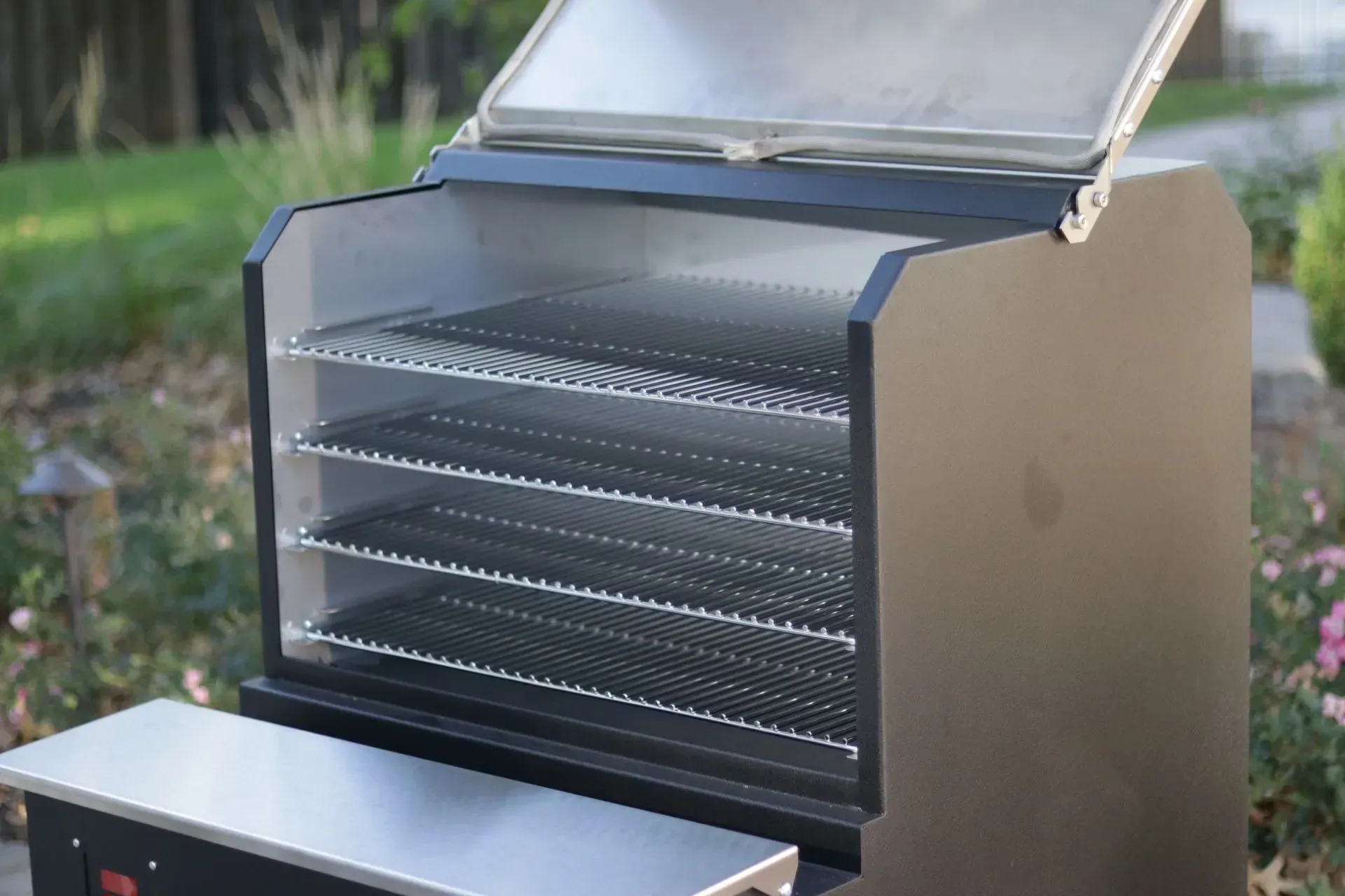 Black smoker grill with multiple wire racks inside and the lid open. Outdoors, with greenery in the background.