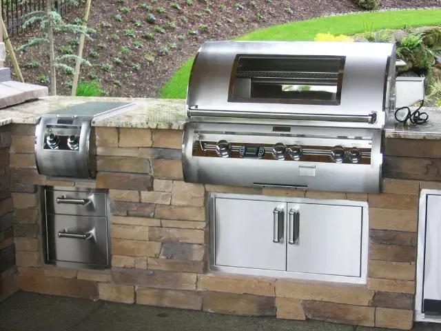 Outdoor kitchen with built-in stainless steel grill, side burner, and storage, set into a stone facade.