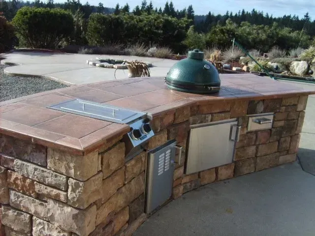 Outdoor kitchen with a stone facade, featuring a grill, Big Green Egg, and stainless steel appliances on a curved countertop.