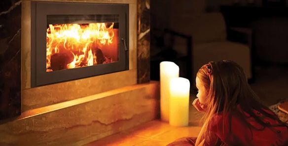 A young girl gazes at a roaring fireplace, illuminated by flickering flames and two large pillar candles in a dimly lit room.