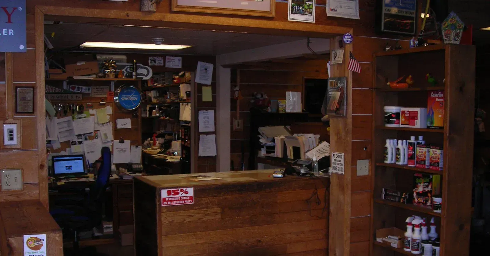 A cluttered wooden counter in a store, likely a general store or shop, with shelves displaying various items and a computer visible.