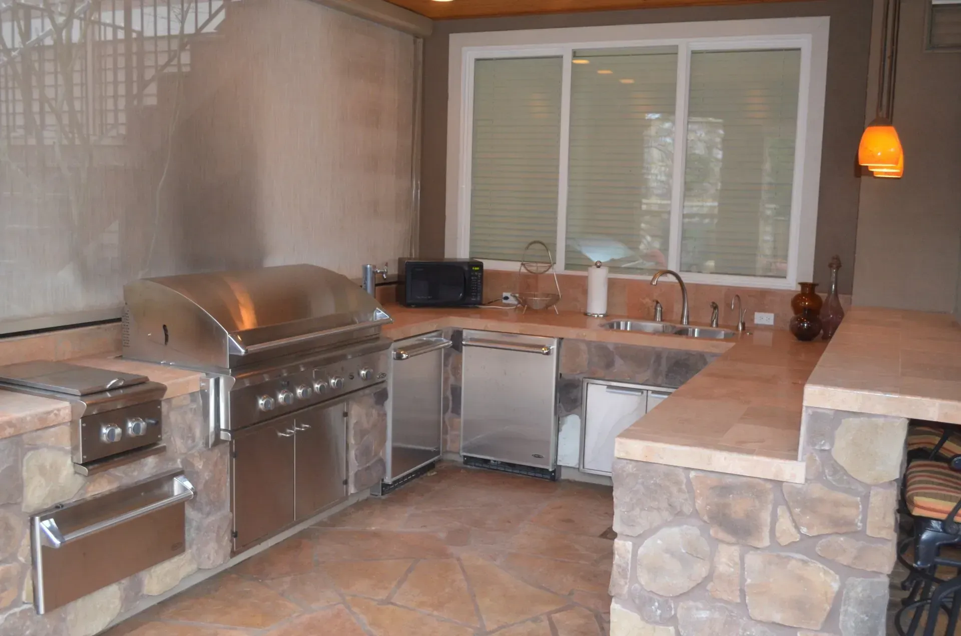 Outdoor kitchen with stainless steel grill and appliances, stone countertops and accents, and a window overlooking the outdoors.