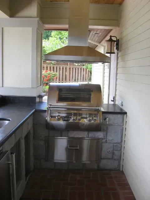 Outdoor kitchen with a stainless steel grill and vent hood built into a stone and brick structure; cream cabinets and black countertops.