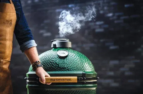 A person opening a green Big Green Egg grill, smoke rising from the top. They wear a leather apron and are outdoors.