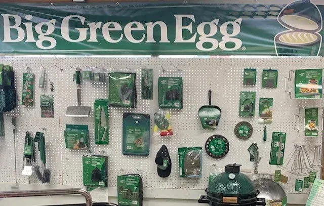 A display of Big Green Egg grilling accessories in a store setting, featuring green and white packaging. The brand's logo is visible on a banner.