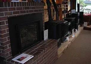 A brick fireplace with a black insert is next to several black stoves on a display in a store.
