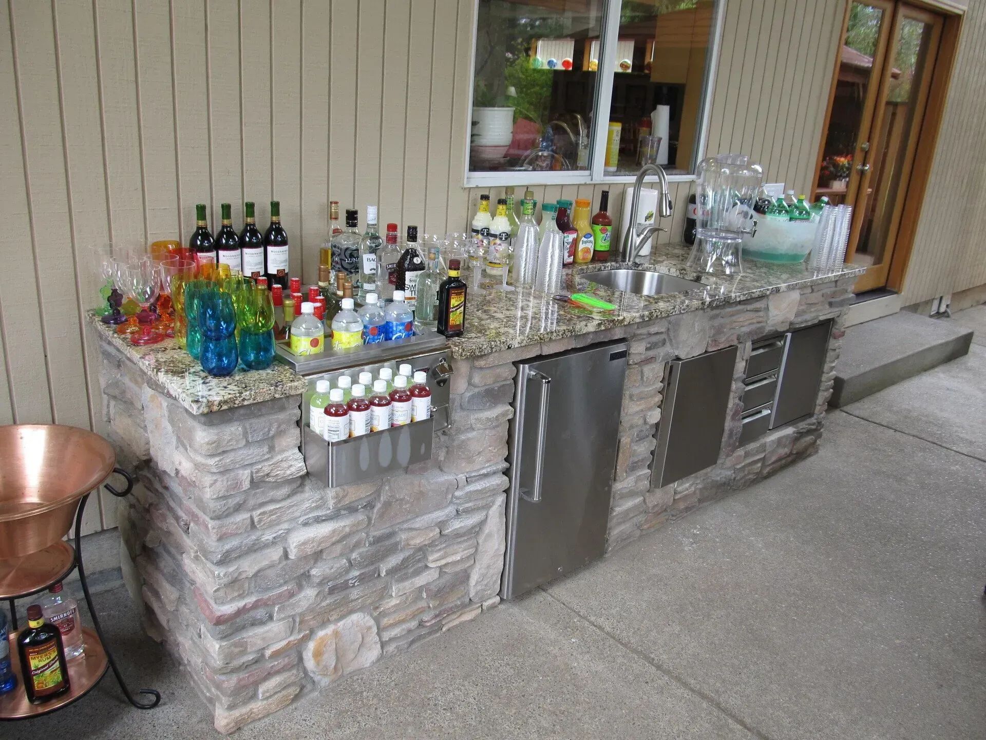 Outdoor bar with a stone facade, granite countertop, and stainless steel appliances. Drinks and supplies are displayed on the counter.