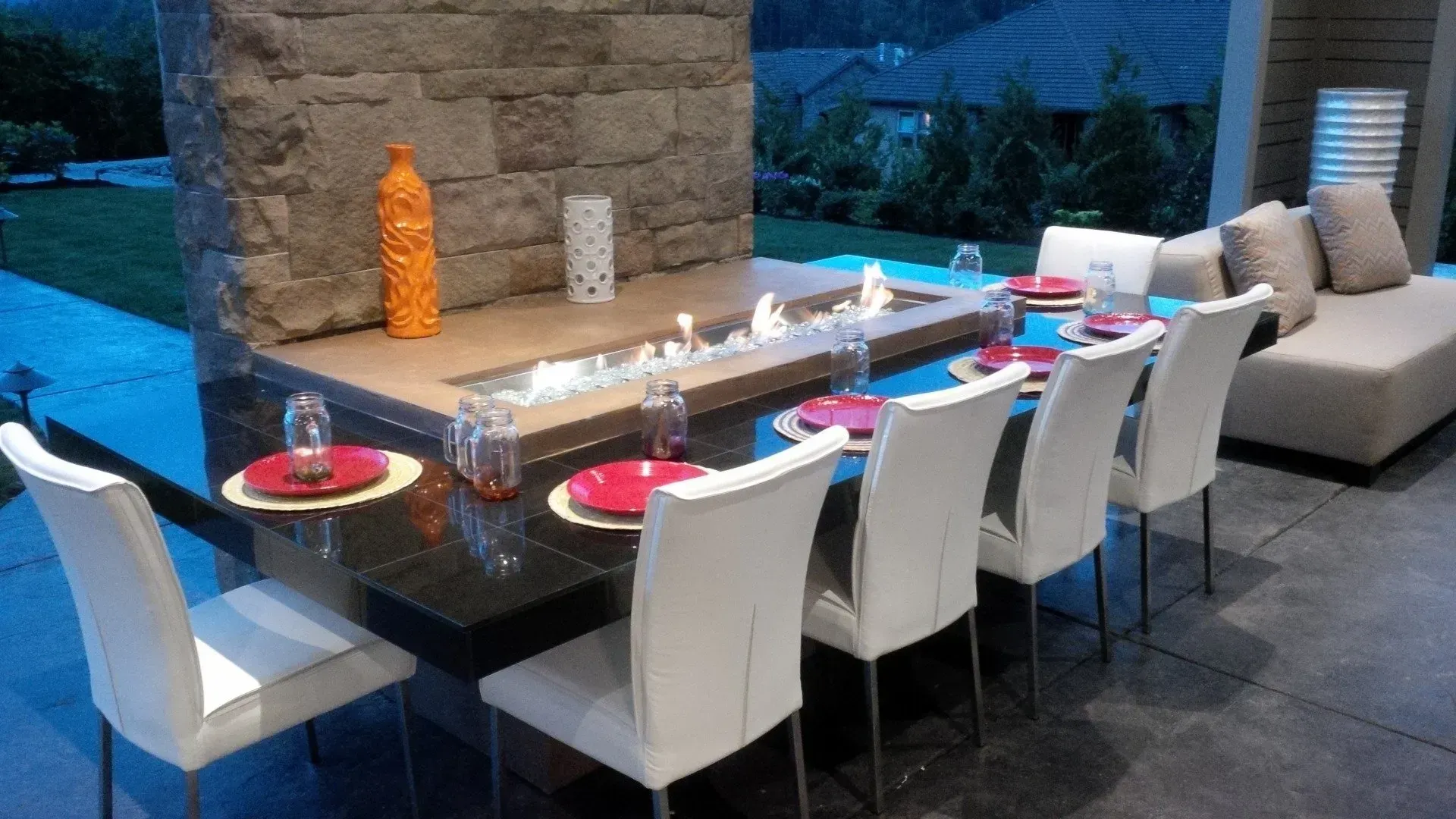 Outdoor dining table set for a meal, with white chairs, red place settings, and a built-in fire feature.