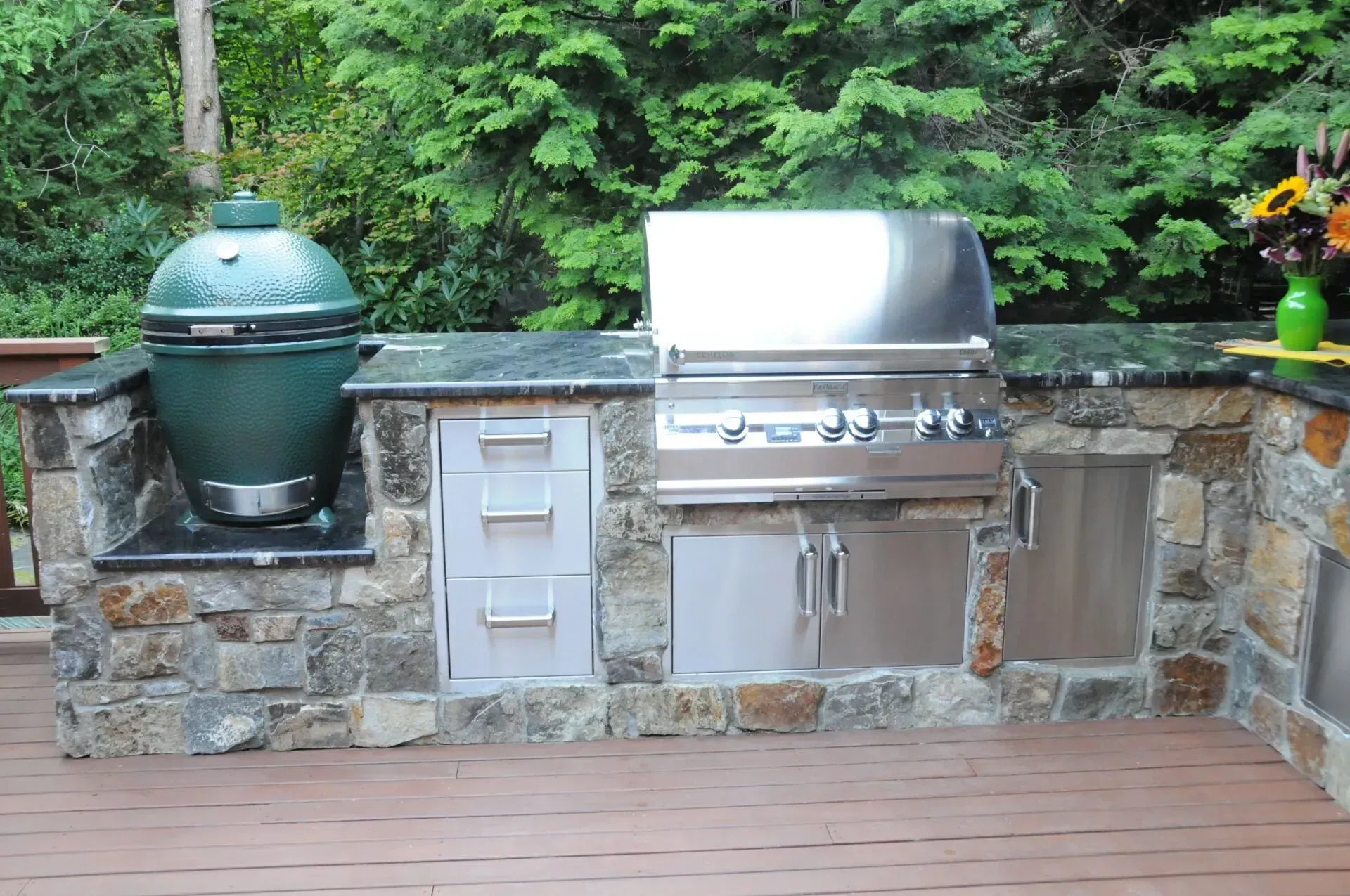 Outdoor kitchen with a green egg grill, stainless steel grill, and stone facade. The setting is a deck with foliage in the background.
