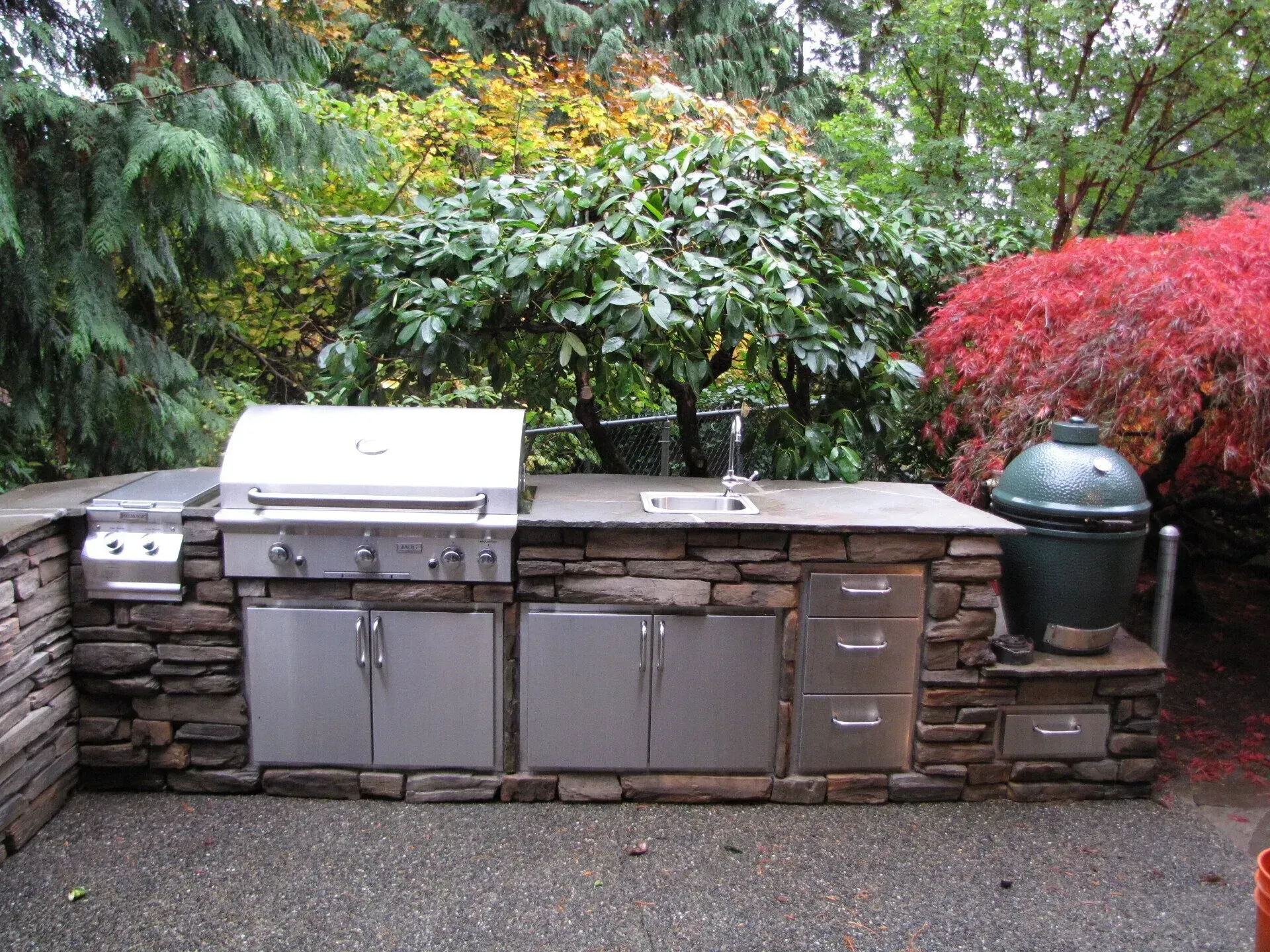 Outdoor kitchen with built-in grill, sink, cabinets, and a green Big Green Egg, set against lush trees.