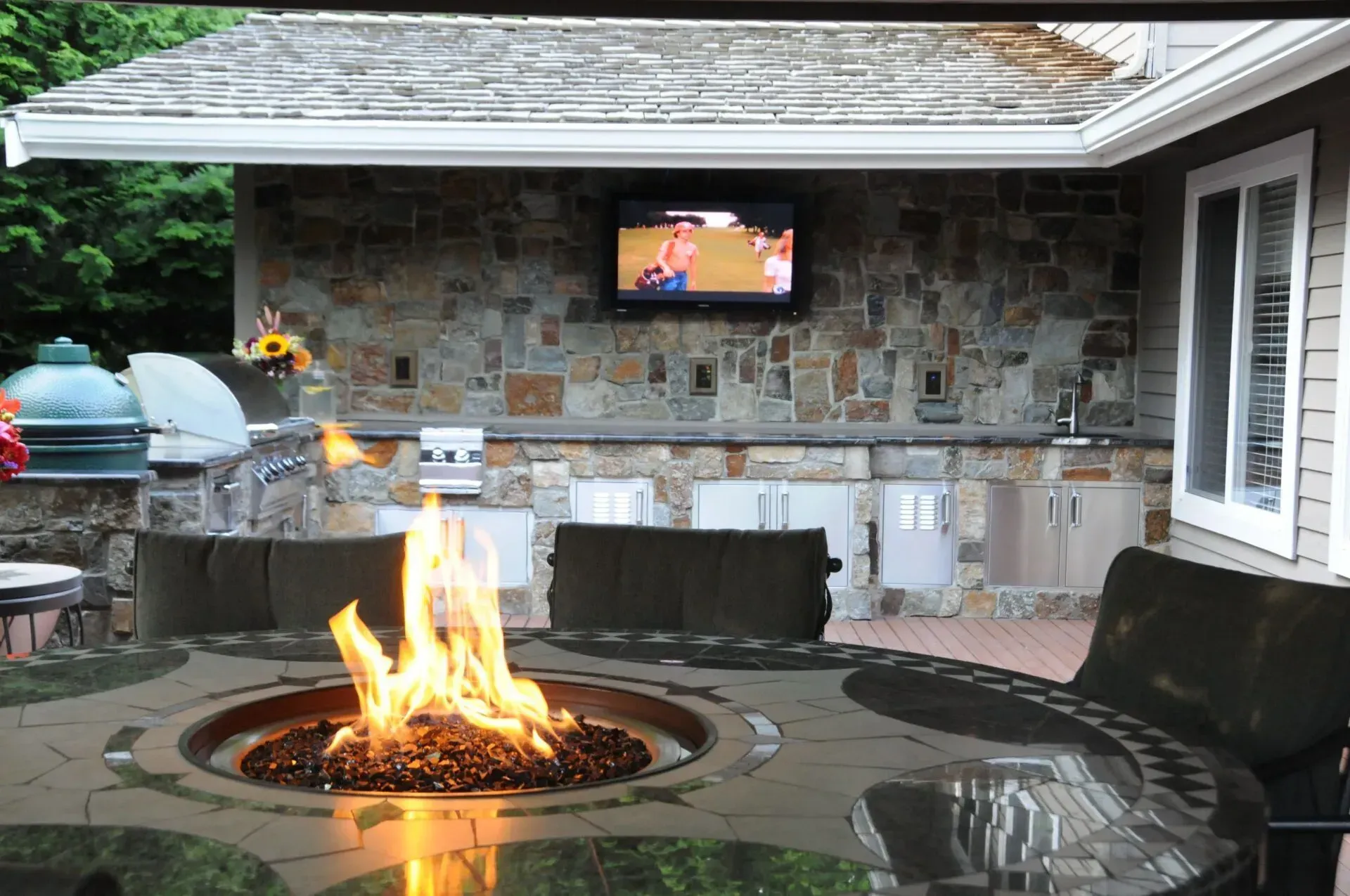 Outdoor kitchen with a lit fire pit and a TV mounted on a stone wall. There's a grill, seating, and cabinets.