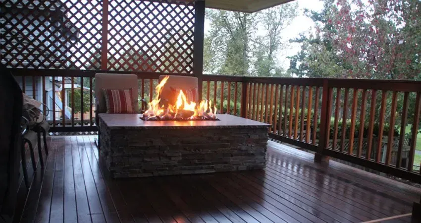 Fire pit on a wooden deck, flames glowing.  Enclosed by a wooden railing and lattice.  Overlooking a yard with trees.
