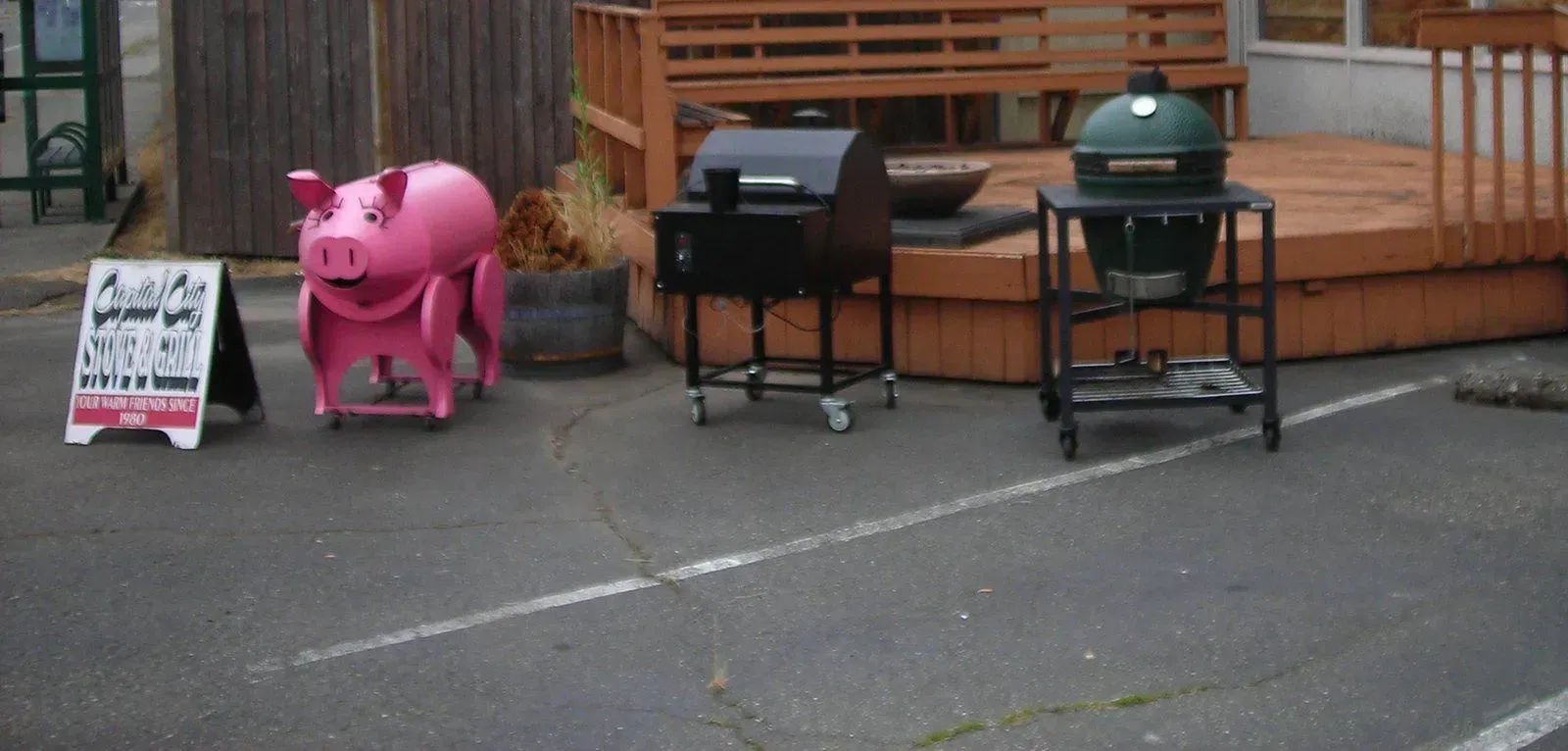 A pink pig-shaped propane tank stands in front of a barbecue equipment store. Other barbecues and a menu sign are also present.
