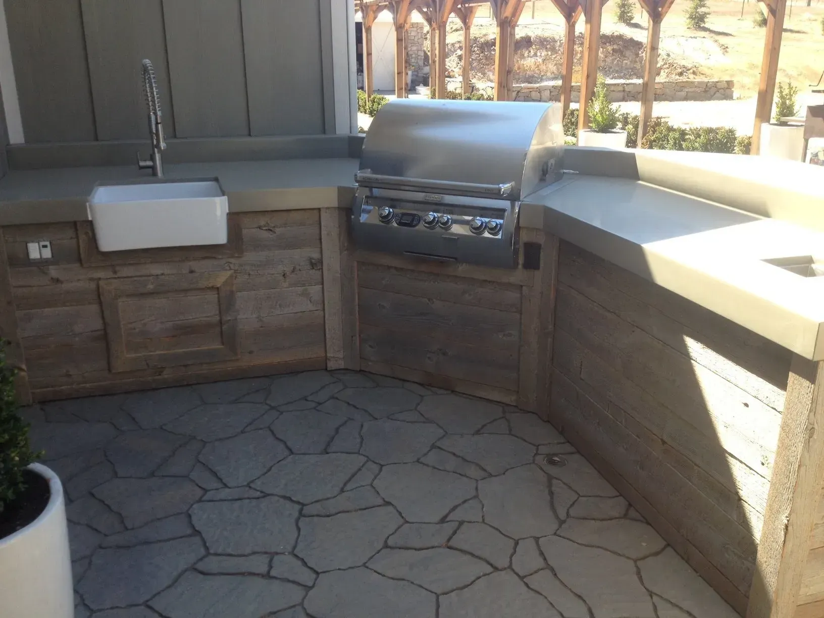 Outdoor kitchen with a stainless steel grill, sink, and stone-like cabinets on a paved patio.