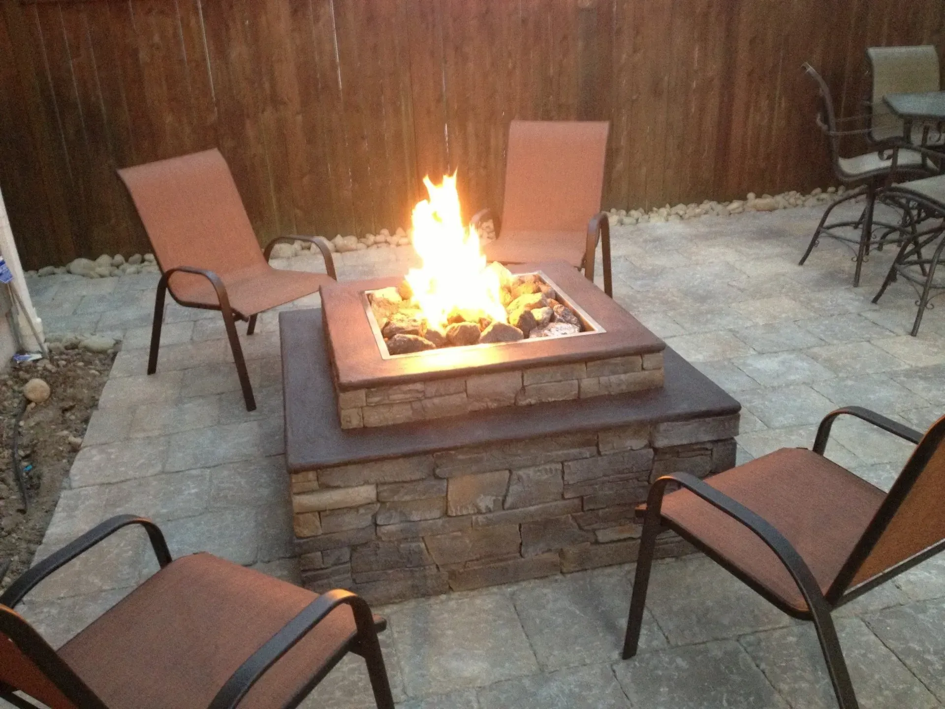 A lit fire pit surrounded by four chairs on a paved patio. Stone and brown tones dominate the scene.