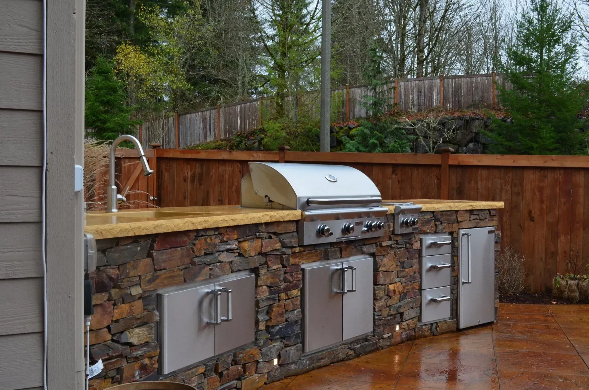 Outdoor kitchen with a stone base, stainless steel appliances, and a wooden fence in the background.