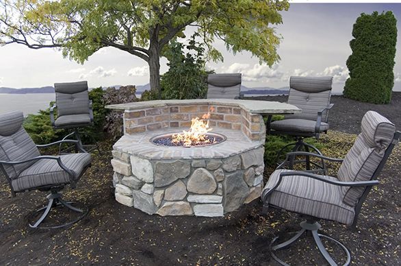 A stone fire pit with a bar is surrounded by four swivel chairs. The setting is outdoors with a view of water.