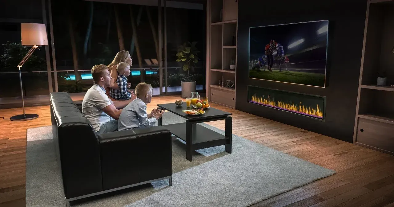 Family watching a football game on a large TV above a fireplace in a modern living room at night.