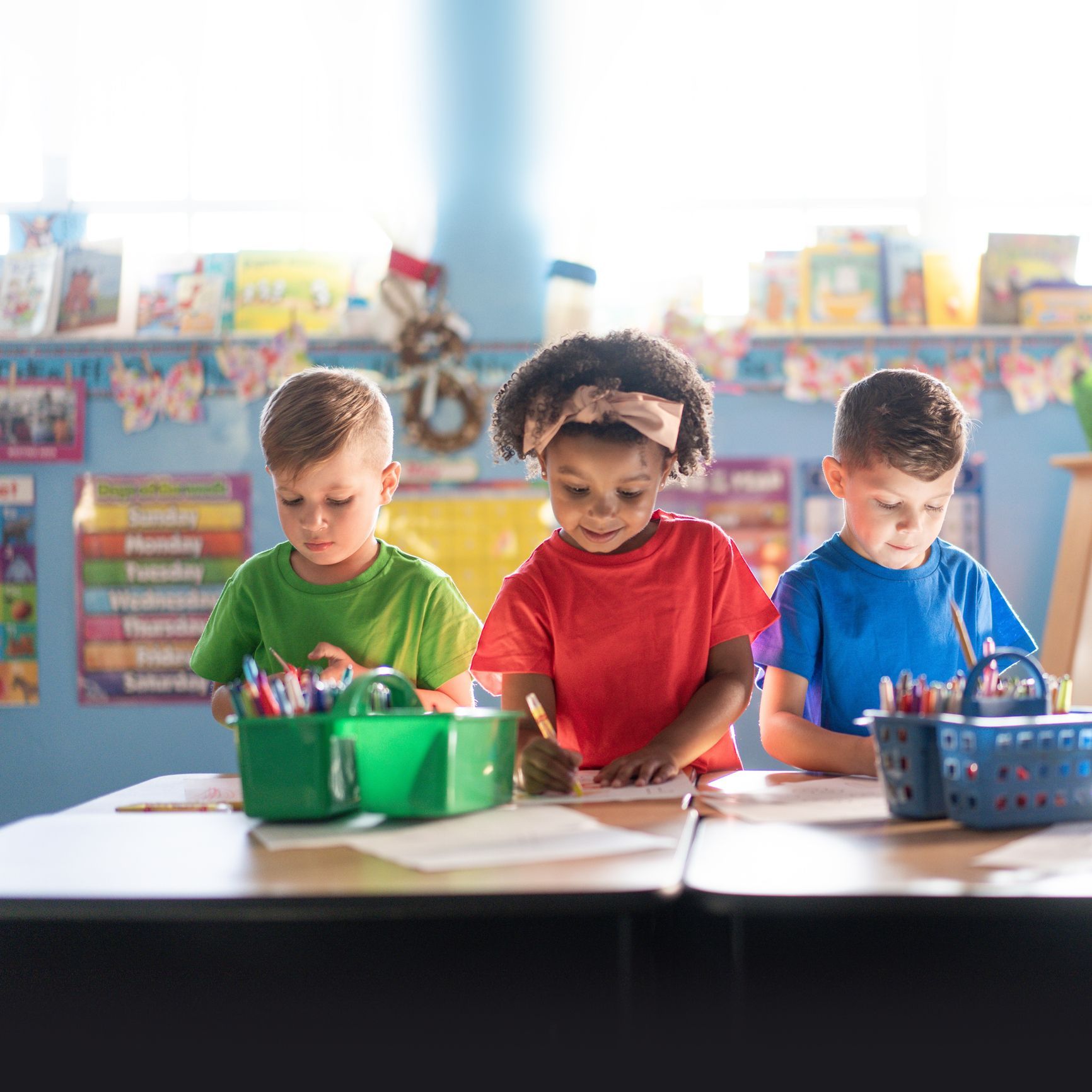 Three young children in a classroom, drawing at a table with crayons and a green basket of pencils.